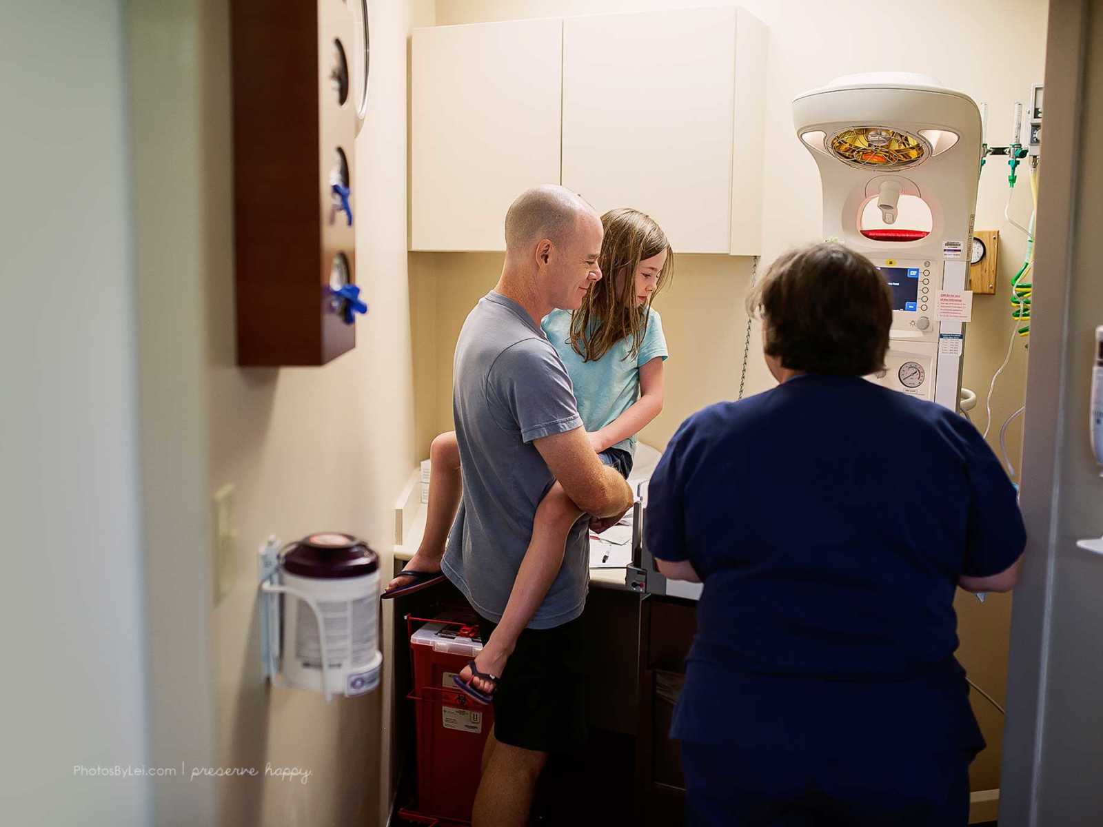 Father of newborn covered in amniotic fluid holds older daughter next to machine with nurse