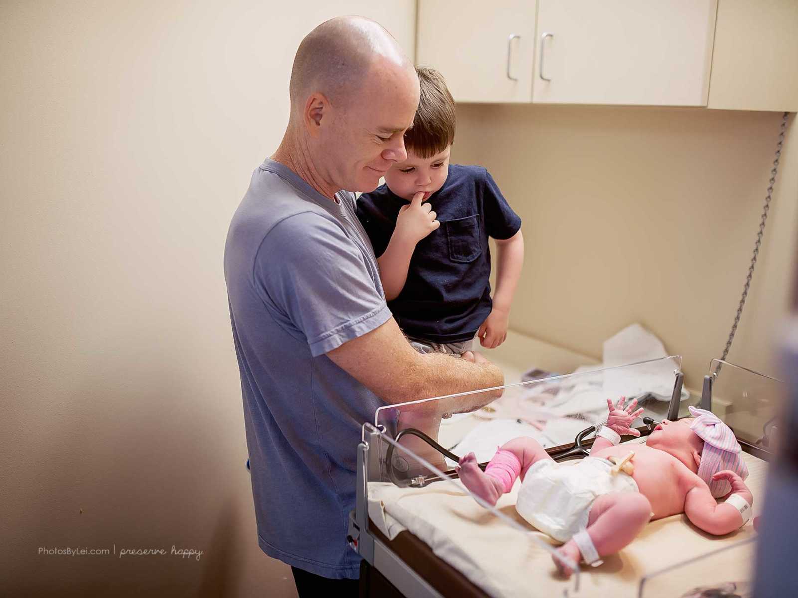 Father of newborn covered in amniotic fluid holds son to look at his new sibling
