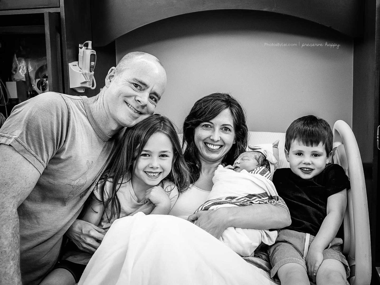 Mother who gave birth to baby covered in amniotic fluid smiles in hospital bed with newborn, son, daughter and husband