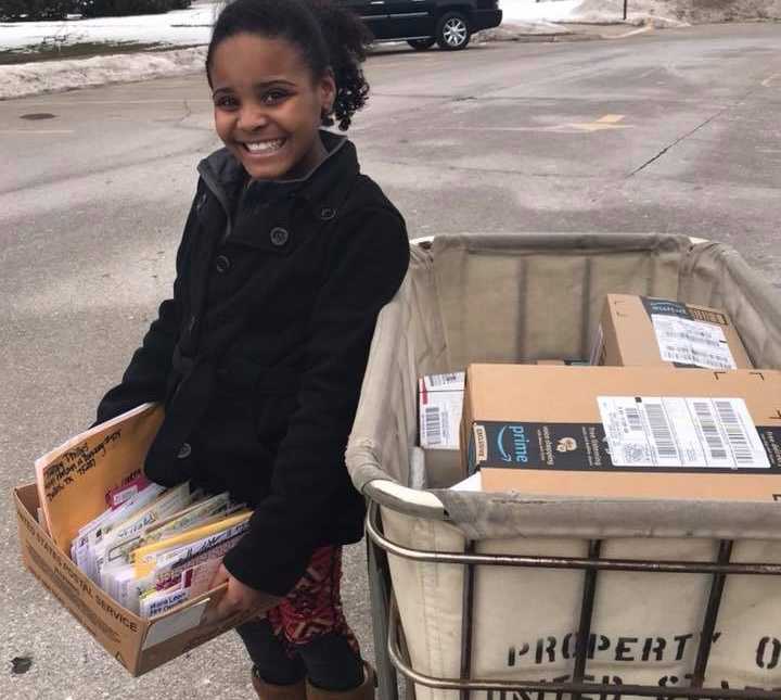 Girl from Flint, Michigan smiles while holding box of letters next to mail card filled with cardboard boxes