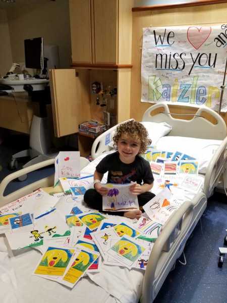 Little boy with leukemia sitting in hospital bed smiling with hand made notes and poster behind him