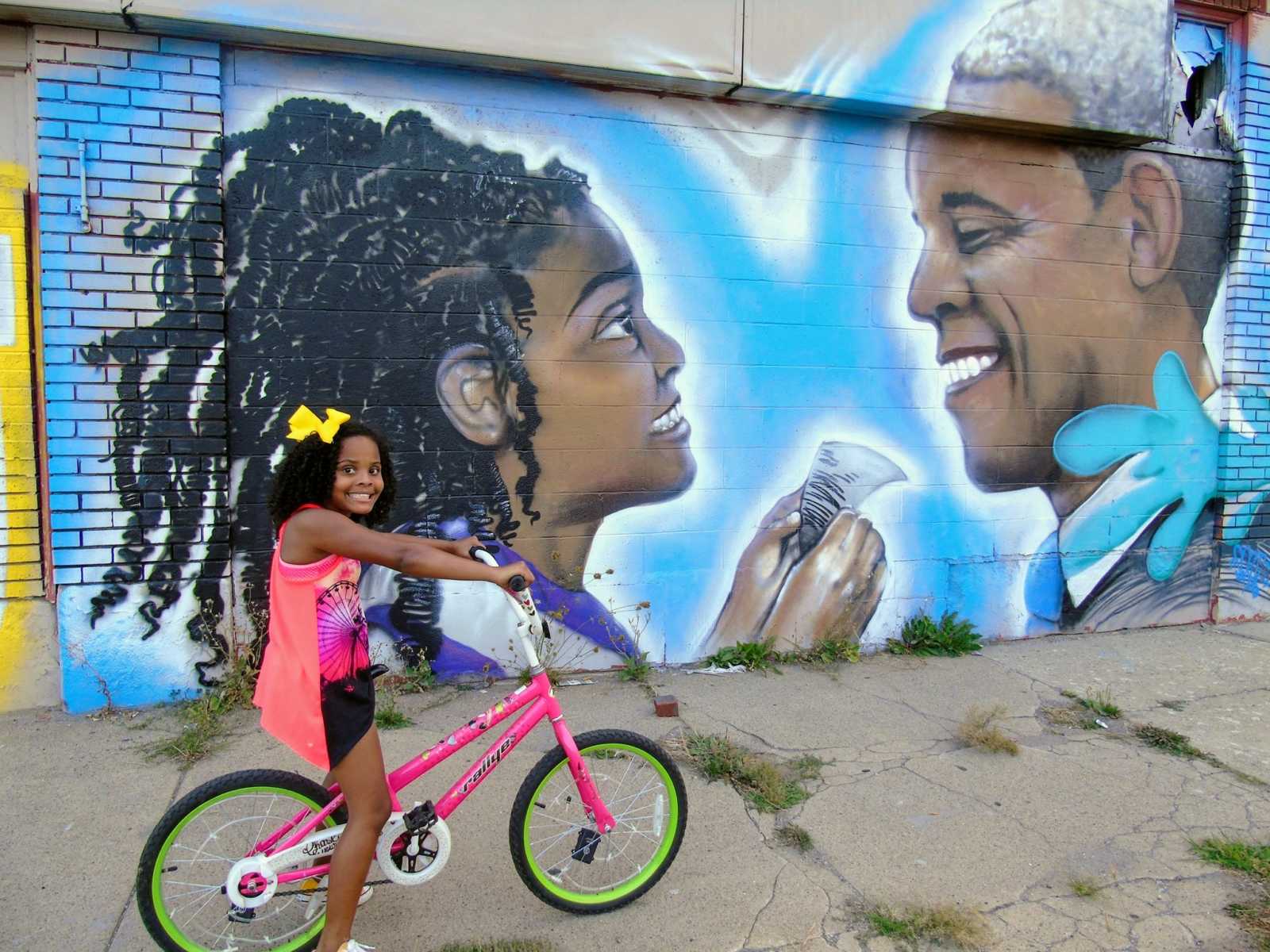 Little girl in Flint, Michigan smiling on her bike in front of mural of a girl handing something to Barack Obama