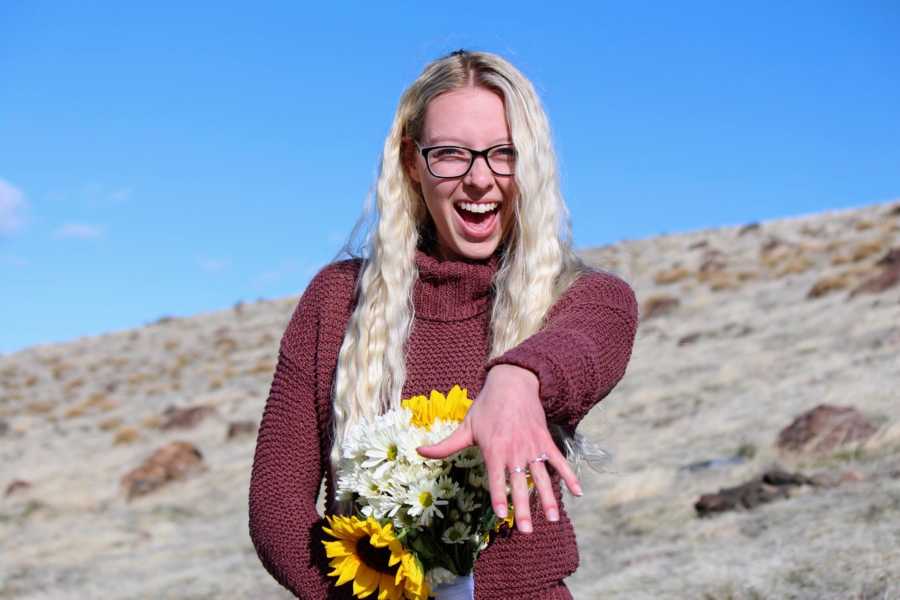 Woman smiles while holding bouquet of flowers in one hand while holding out other hand with engagement ring