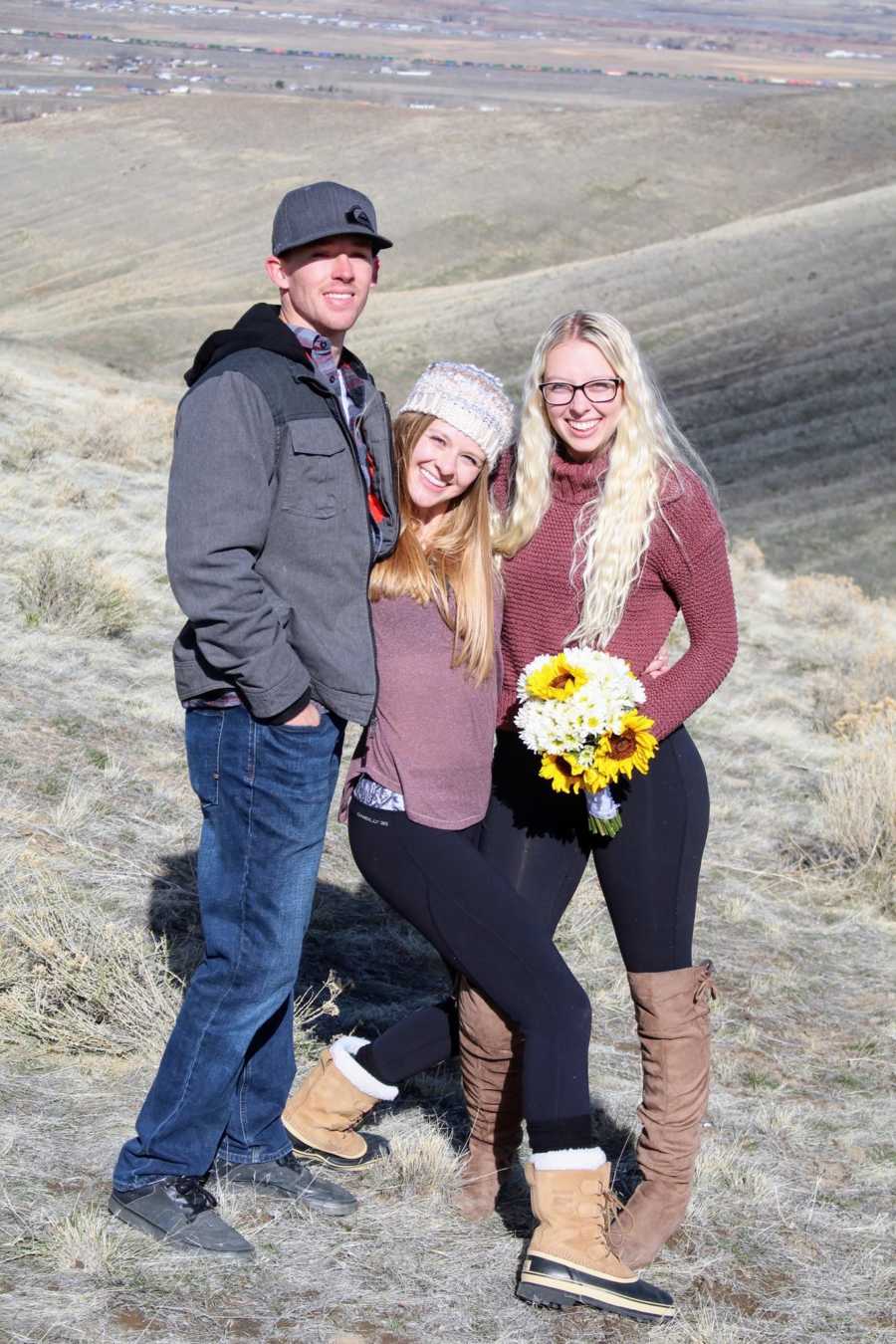 Woman who was just proposed to stands with bouquet of flowers with sister and fiancee