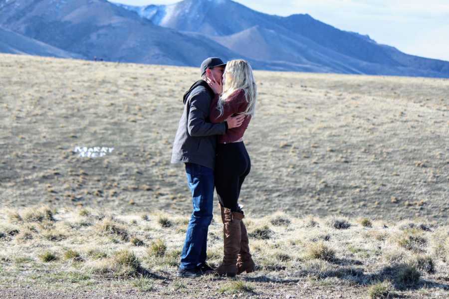 Man and woman kiss after proposal with words, "Marry me" on ground in background