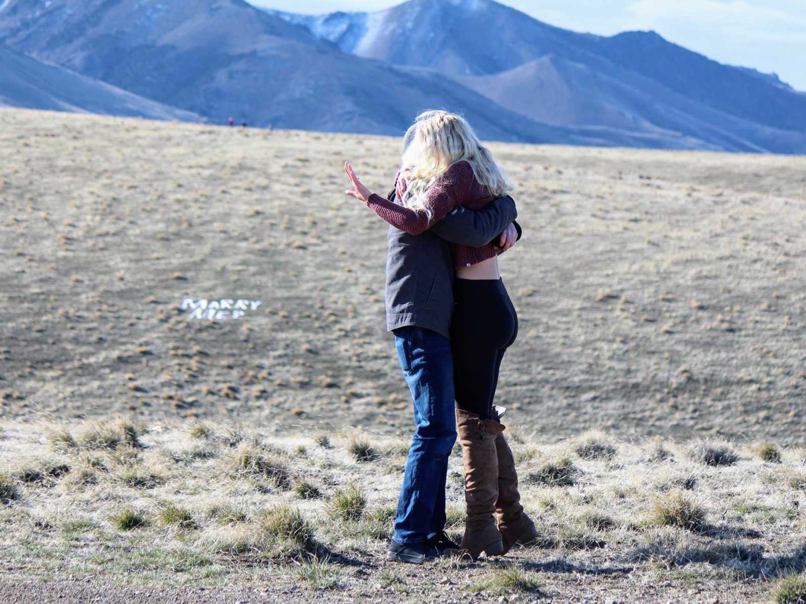 Man and woman hug as woman looks at engagement ring with words, "marry me" written on ground