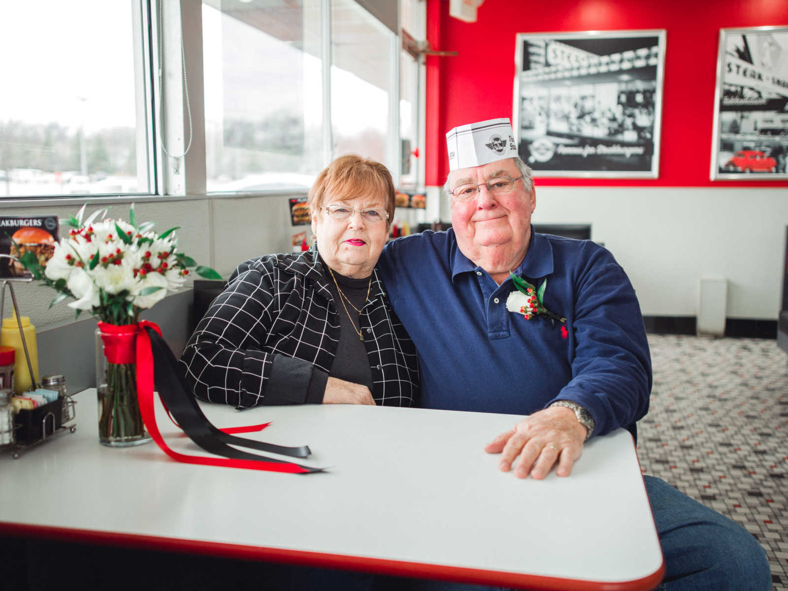 Couple who met at Steak 'n Shake in 1962 smile in booth while husband wears paper hat