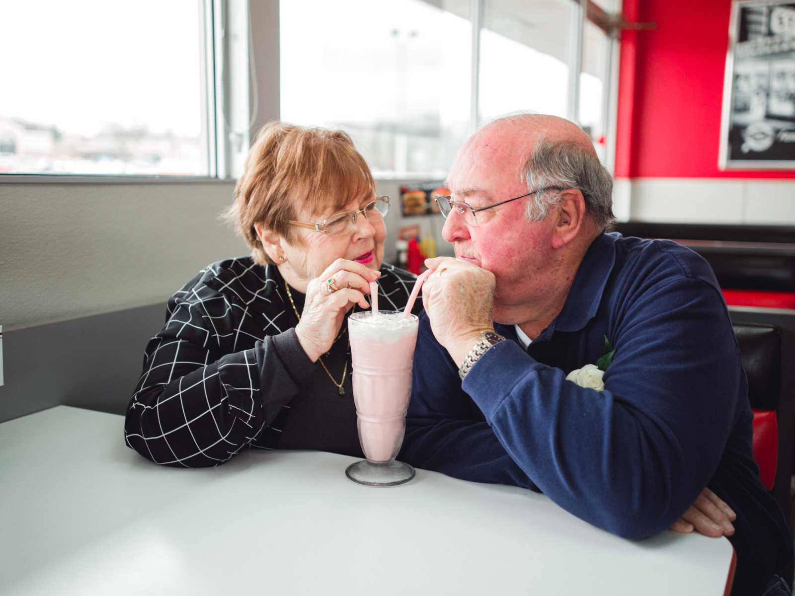 Elderly couple sit in same side of booth in steak 'n shake sharing a milkshake