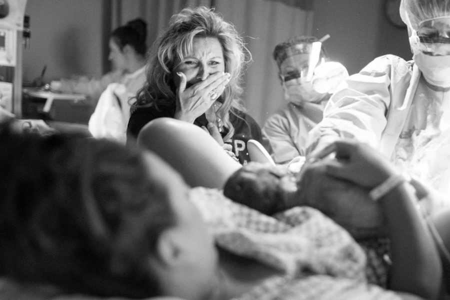 Mother holds hand to mouth crying as she looks at daughter holding her newborn