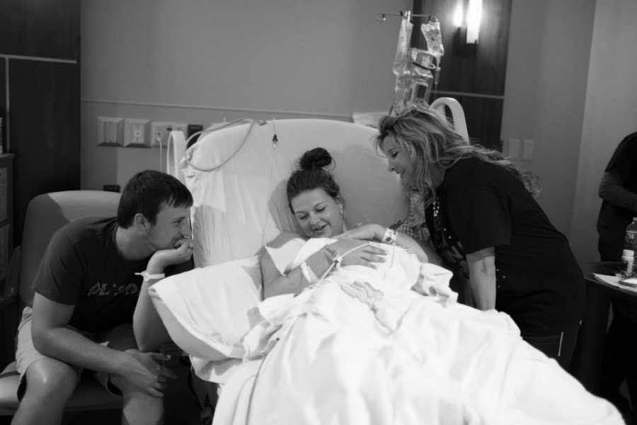 Woman who just gave birth in hospital bed holds newborn with her mother and husband watching