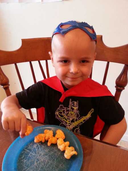Boy with leukemia sitting at table eating food while wearing red cape and blue eye mask on his head