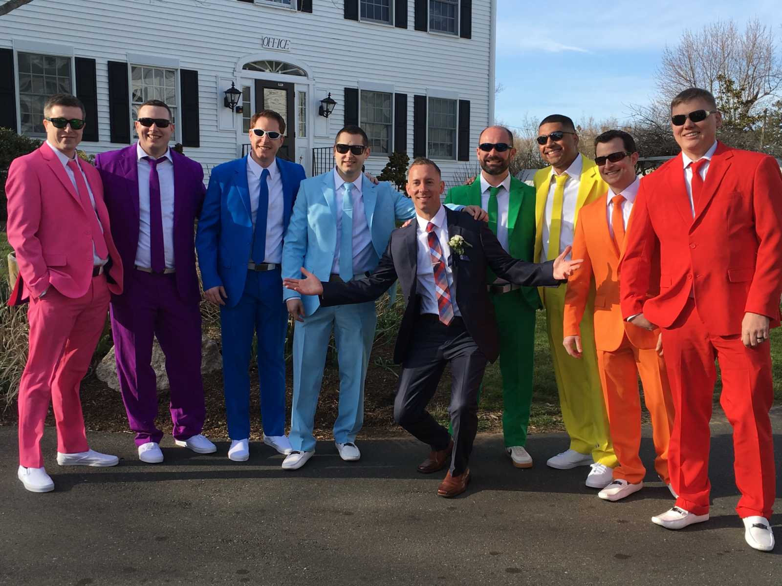 Groom squats and poses with friends in rainbow colored suits smile on either side of him