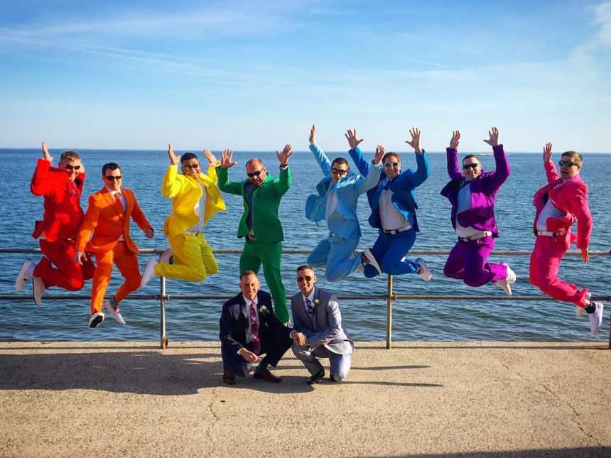 Grooms kneel on pier while friends in suits in colors of rainbow jump in the air behind them