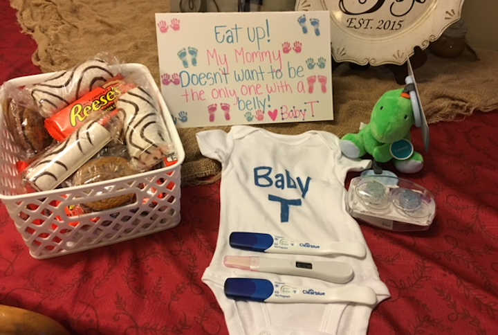 Baby onesie with pregnancy tests on it next to sign and stuffed animal and basket of candy