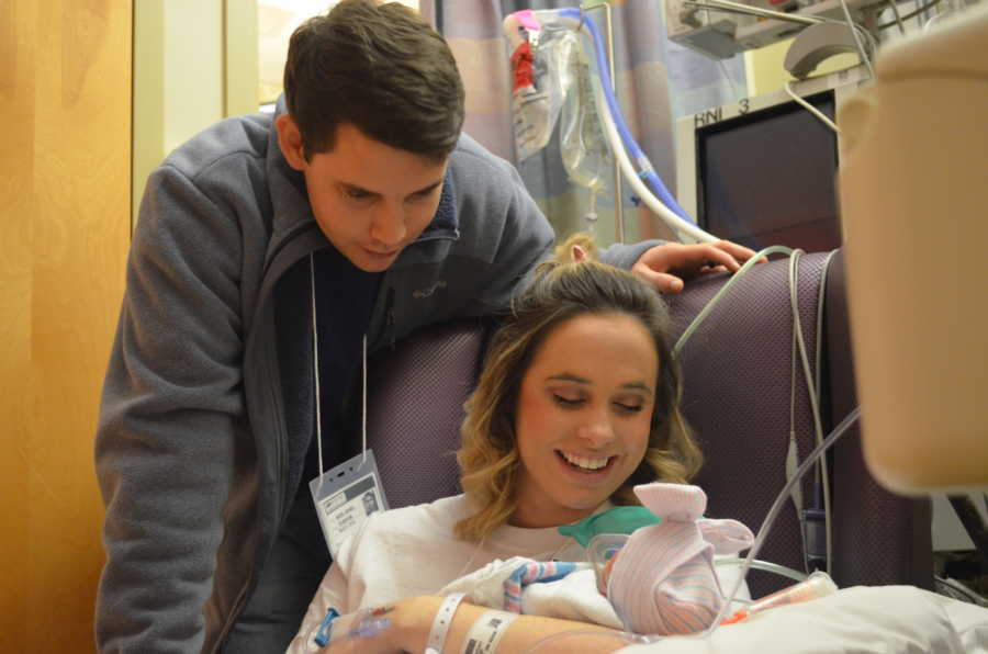 Mother smiles down at two pound newborn in her arms with husband looking at baby over wife's shoulder