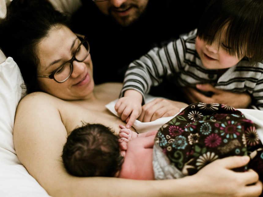 Mother looks down at newborn breastfeeding while firstborn touches baby sister's hand