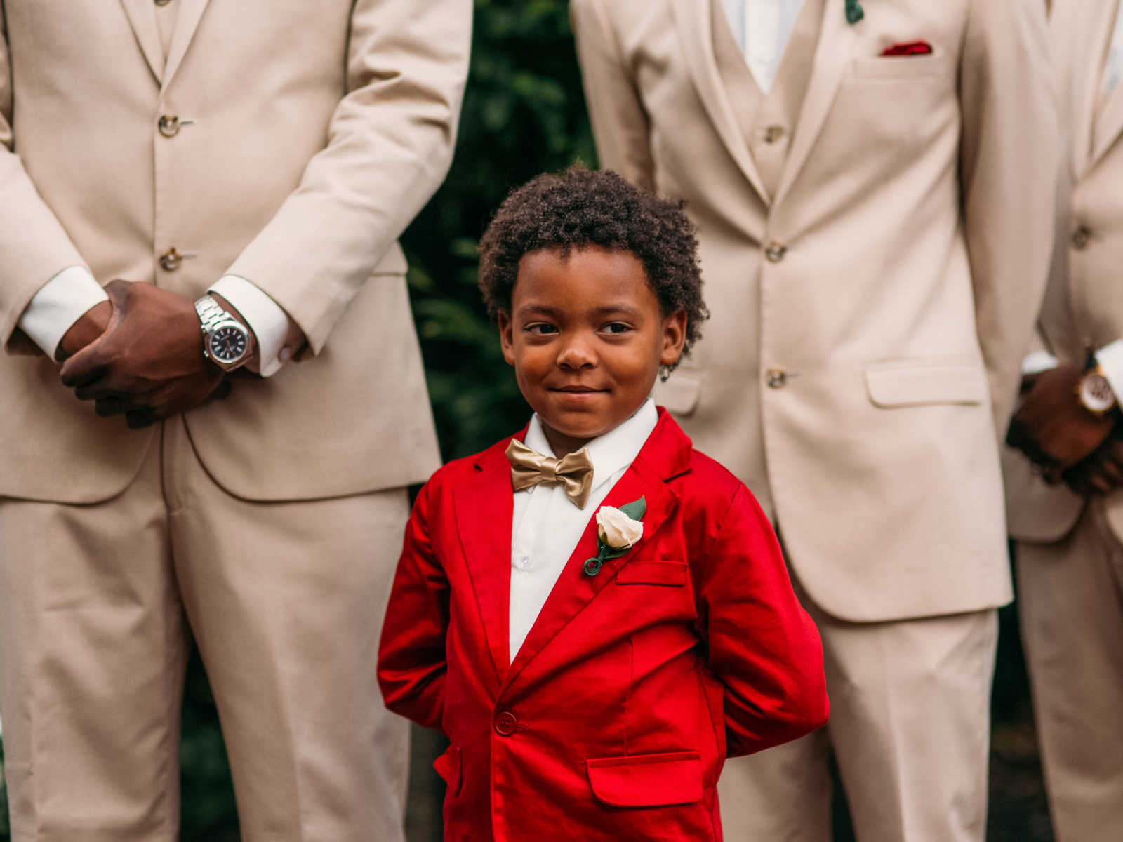 Son of bride smiles in red suit with hands behind his back