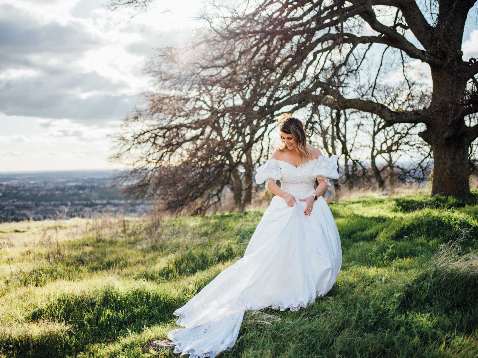 Daughter wearing deceased mother's wedding gown looks down to the side smiling in front of large tree