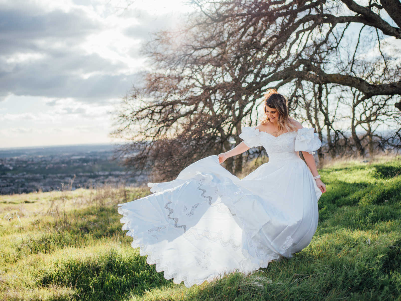 Daughter looks down smiling while holding out skirt of late mothers wedding dress in field