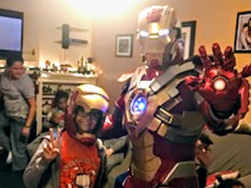 Little boy with cancer wearing iron man helmet holds uo peace sign while standing next to ironman in his home