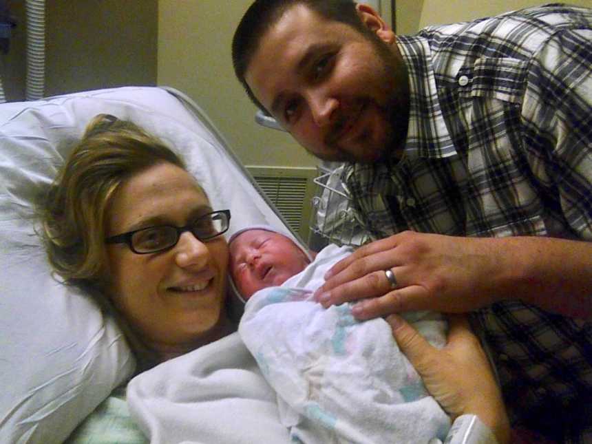 Mother who just gave birth lies in hospital bed holding newborn with her husband at her side