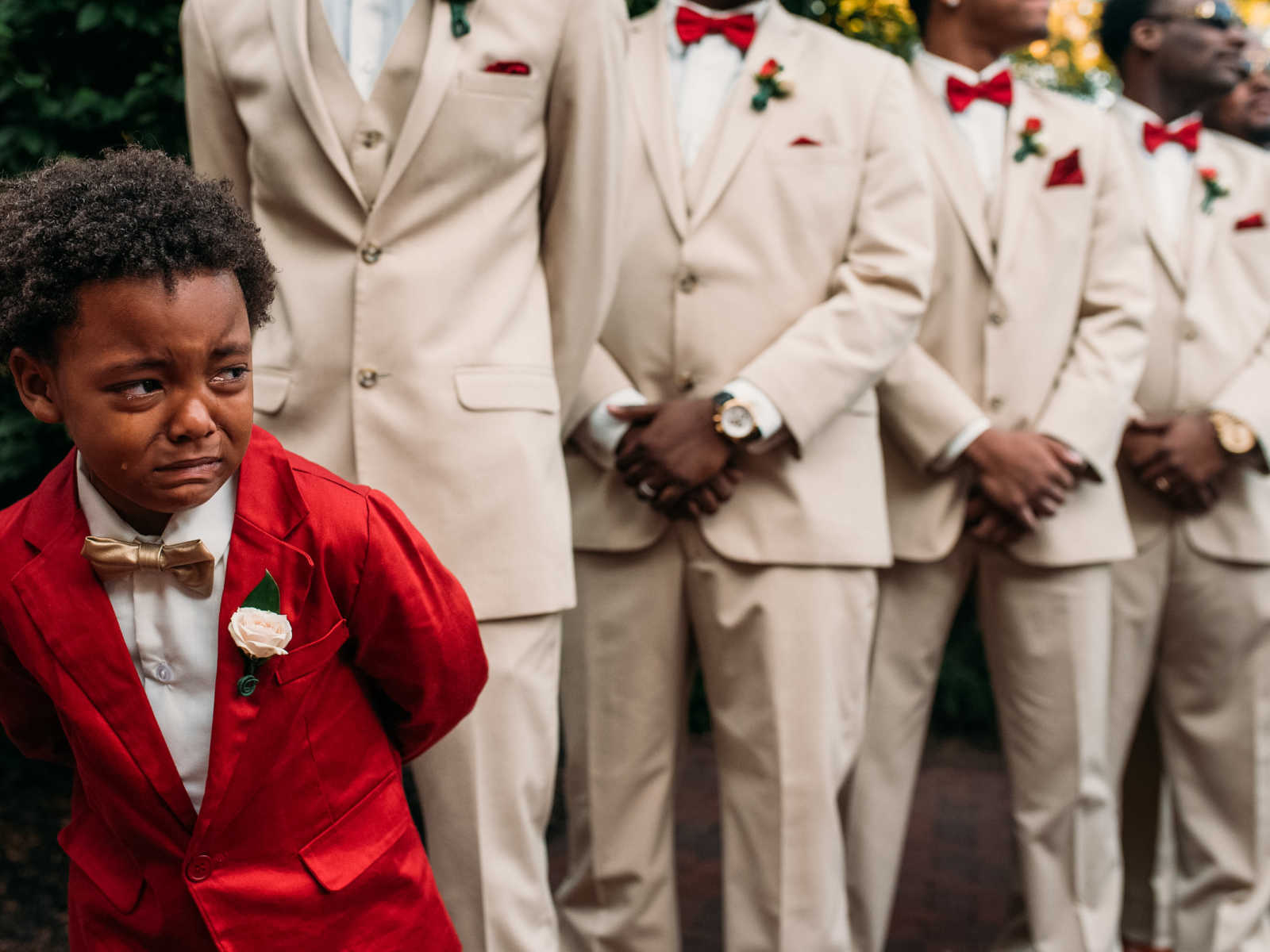 Little boy in red suit stands in front of groomsmen crying as his mother walks down the aisle