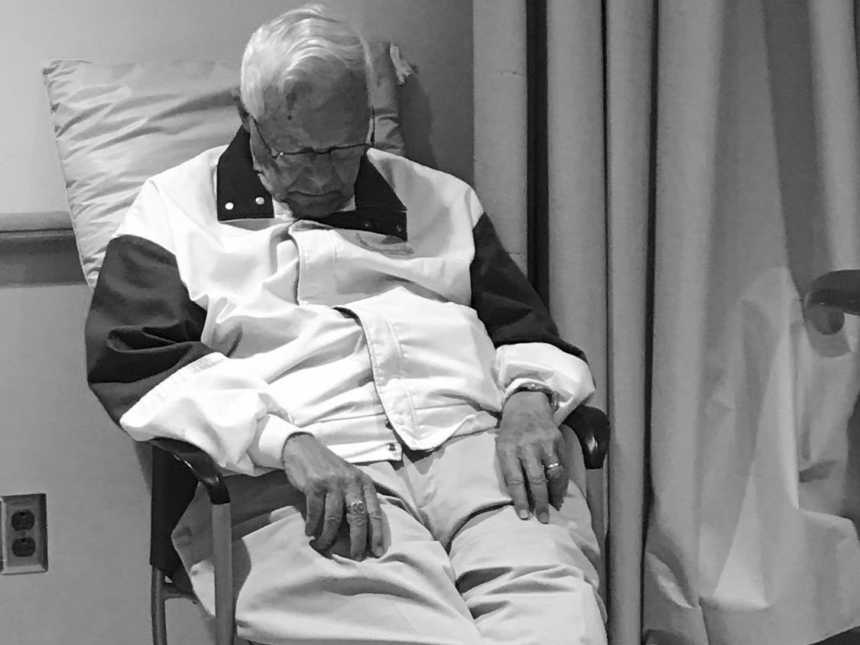 Older man sits hospital chair sleeping with pillow supporting his back