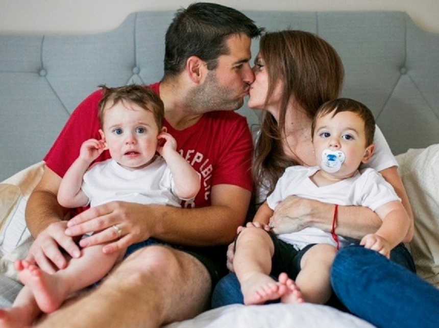 Husband and wife kiss in bed with toddler twins in their lap which were conceived through IVF