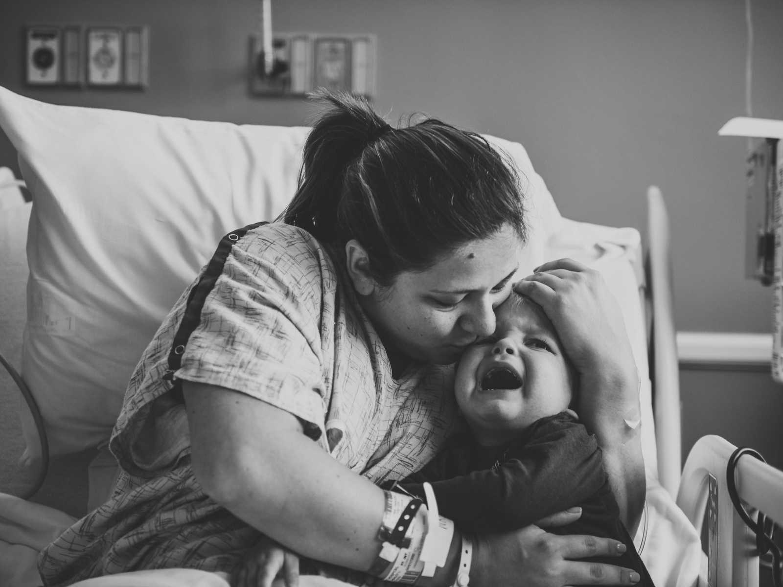 Mother sits up in hospital with arms wrapped around first born who is crying