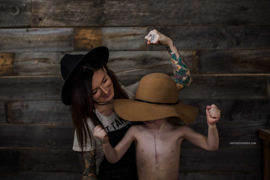 Mother stand smiling behind her son who has had three heart surgeries as he flexes his muscles 