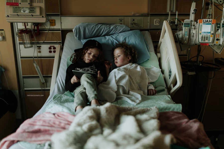 Four year old who just had third heart surgery sits in hospital bed with older brother watching something on tablet