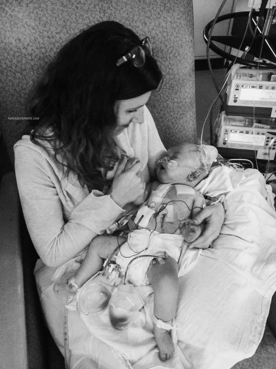 Mother smiles down at baby sleeping with hospital machines hooked up to him after second heart surgery