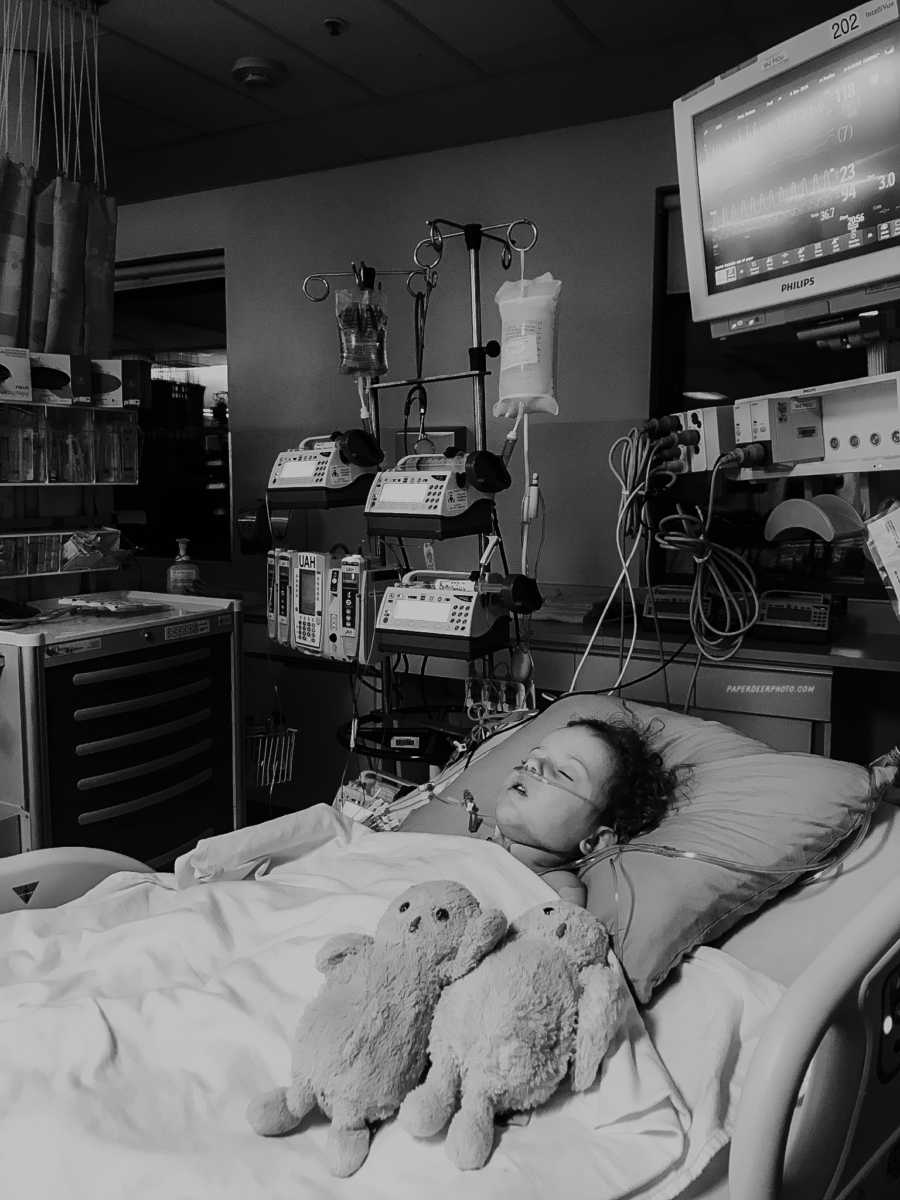 Four year old sleeping in hospital bed next to two stuffed animals after third heart surgery with machines behind him