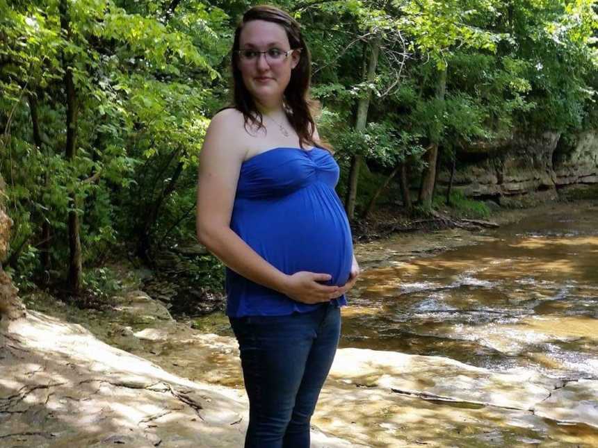 Pregnant woman who was once an addict stands on rock near river holding her stomach