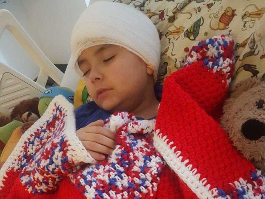 Little boy with cancer sleeps in hospital bed with bandage wrapped around his head wrapped in blacker
