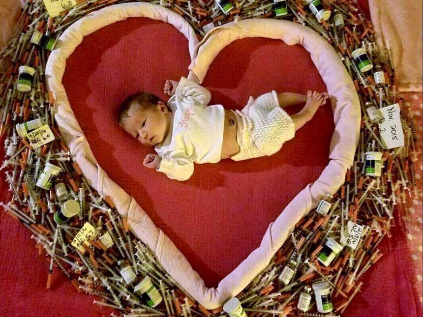Newborn lies on red blanket enclosed by a blanket formed in the shape of a heart with syringes surrounding it