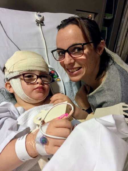 Mom leans over hospital bed smiling next to daughter who just had brain surgery