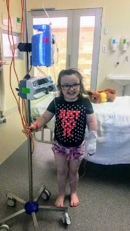 Little girl smiling while standing next to a hospital machine with case on her arm