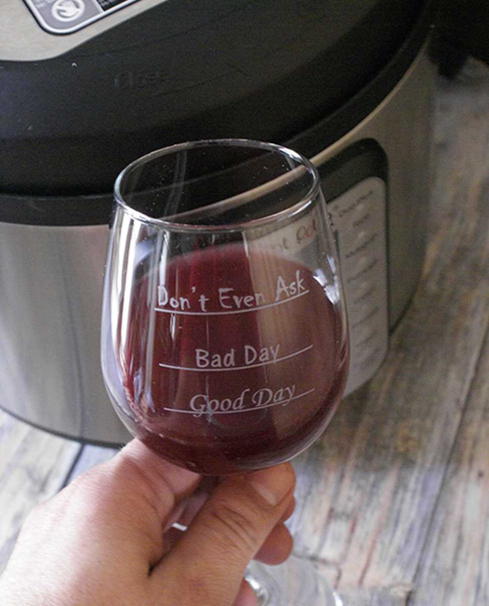 Hand holding up glass of red wine that has lines that say, "don't even ask, bad day, good day" in front of instant pot