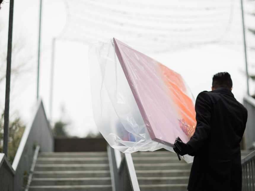 Man walking upstairs holding pink and orange painted canvas in cellophane terminally ill bride's wedding