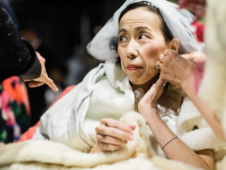 Terminally ill bride with veil on looks to the side as someone puts on her earrings