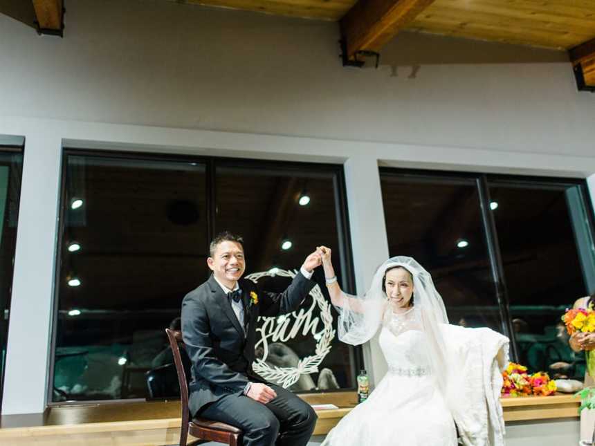 Groom and terminally ill bride sit in chairs smiling while holding hands in the air