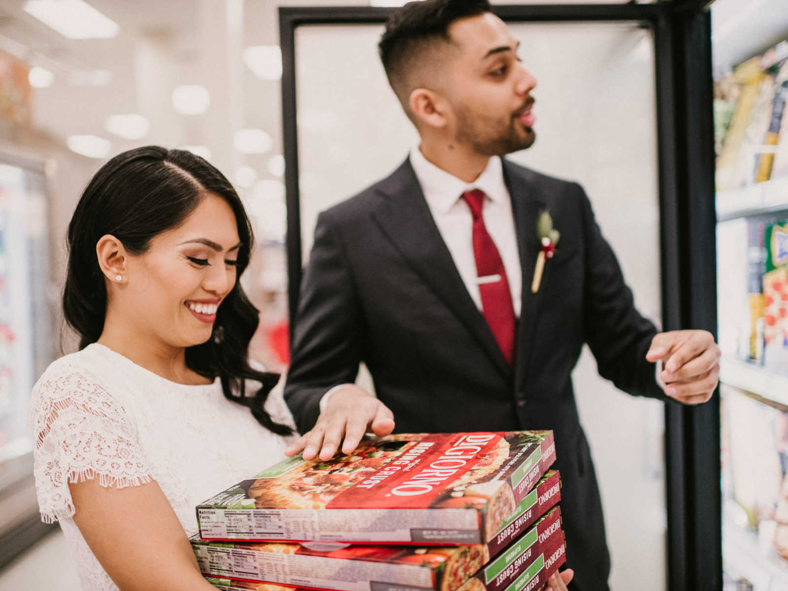Bride smiles down at stack of frozen pizza in her hands while groom adds to it at Target