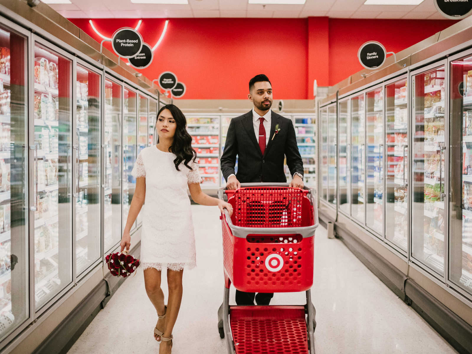 Groom pushes Target shopping cart down freezer aisle while bride holds on to side of cart