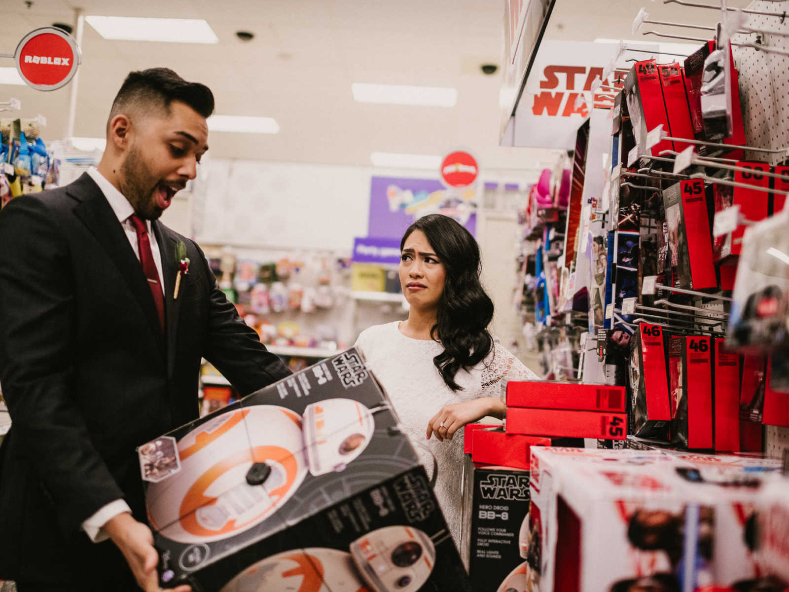 Bride looks at groom with puzzled look as he holds up Star Wars toy in excitement in Target aisle
