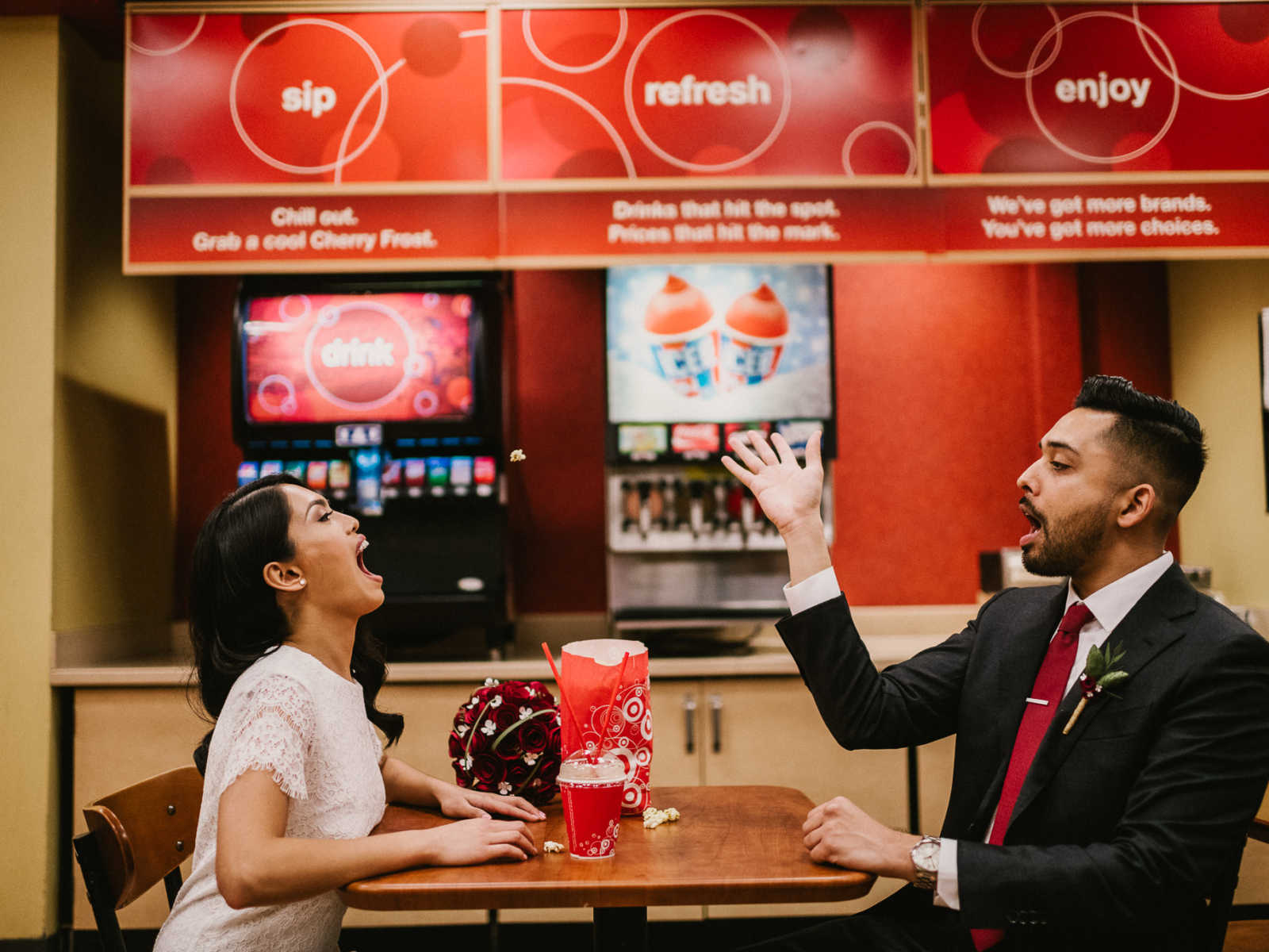 Groom throws popcorn in air for bride to catch in her mouth as they sit at table in Target
