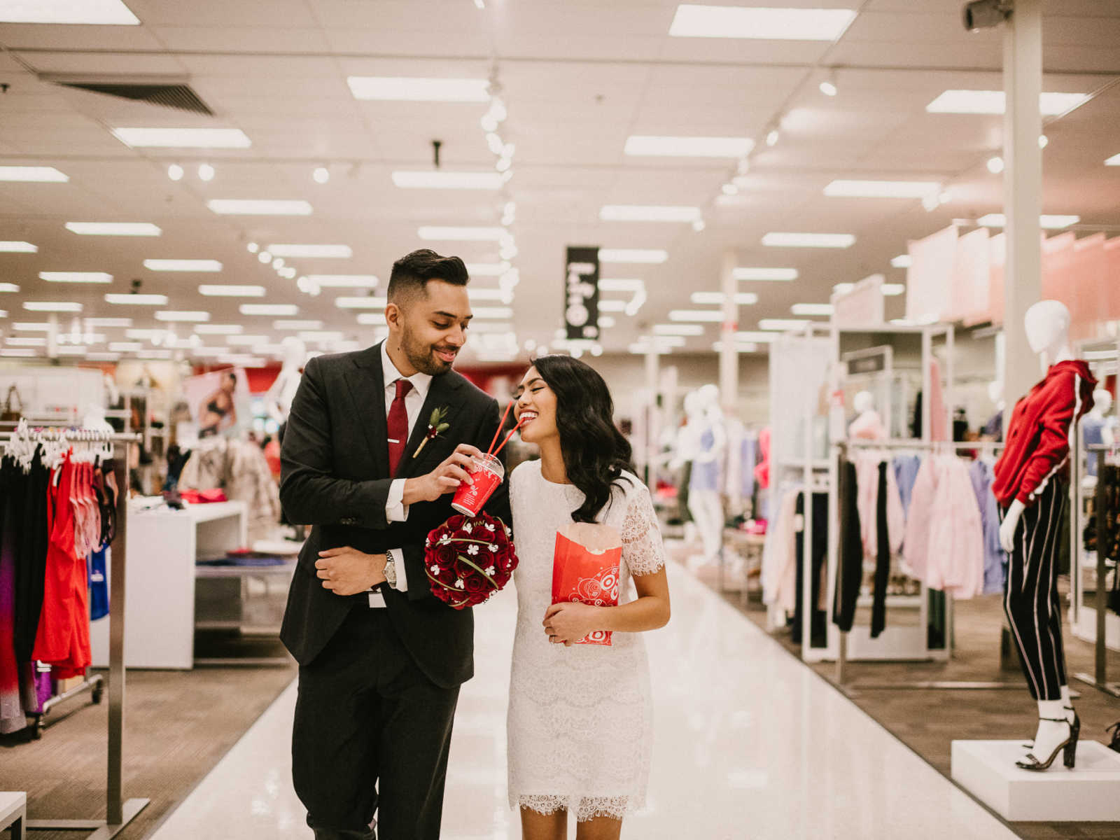 Bride and groom walk arm in arm down Target aisle while groom feeds drink to bride who is holding popcorn bag