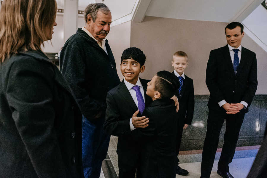 Adopted sons hug each other while mother, father, new brother, and man watch