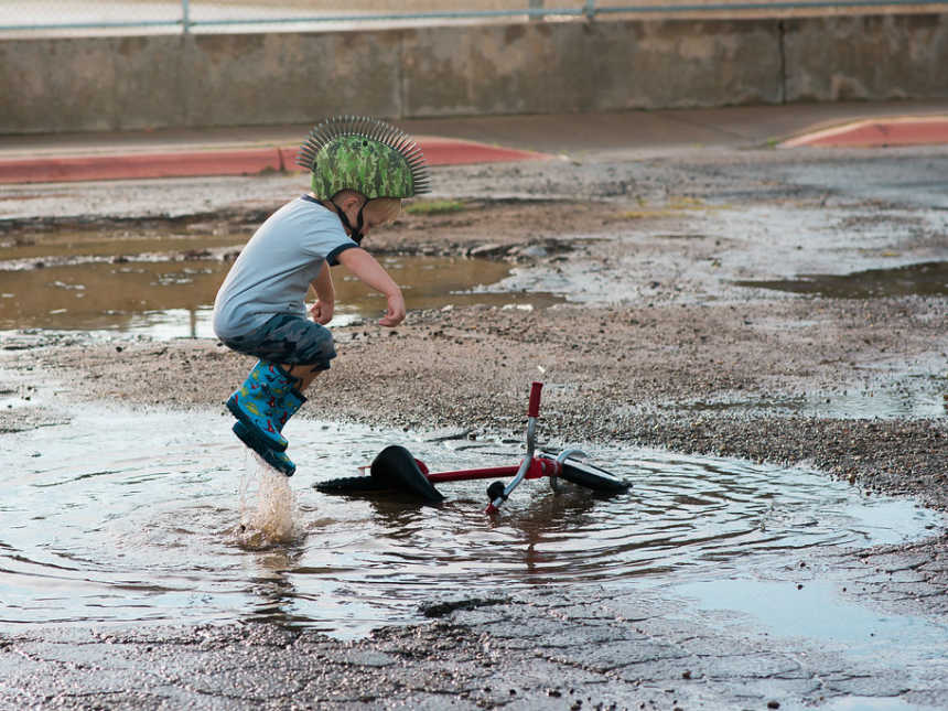 Little boy in mohawk helmet jumping in a puddle where his bike is lying 