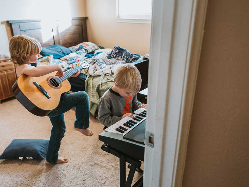 little boy sits in bedroom playing piano while older brother stands behind playing guitar